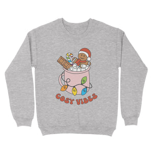 Heather Grey sweatshirt featuring retro text reading 'cosy vibes'. The image shows a mug of hot chocolate with marshmallows and a gingerbread man wearing a Santa hat. A candy cane in the mug and lights around the mug are the colours of the rainbow pride flag.