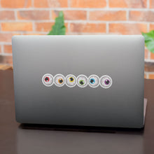 Load image into Gallery viewer, A row of six circular stickers with eyeball designs is aligned on the back of a grey laptop. The irises are a mix of colours including red, orange, yellow, green, blue, and purple, representing the rainbow pride flag.