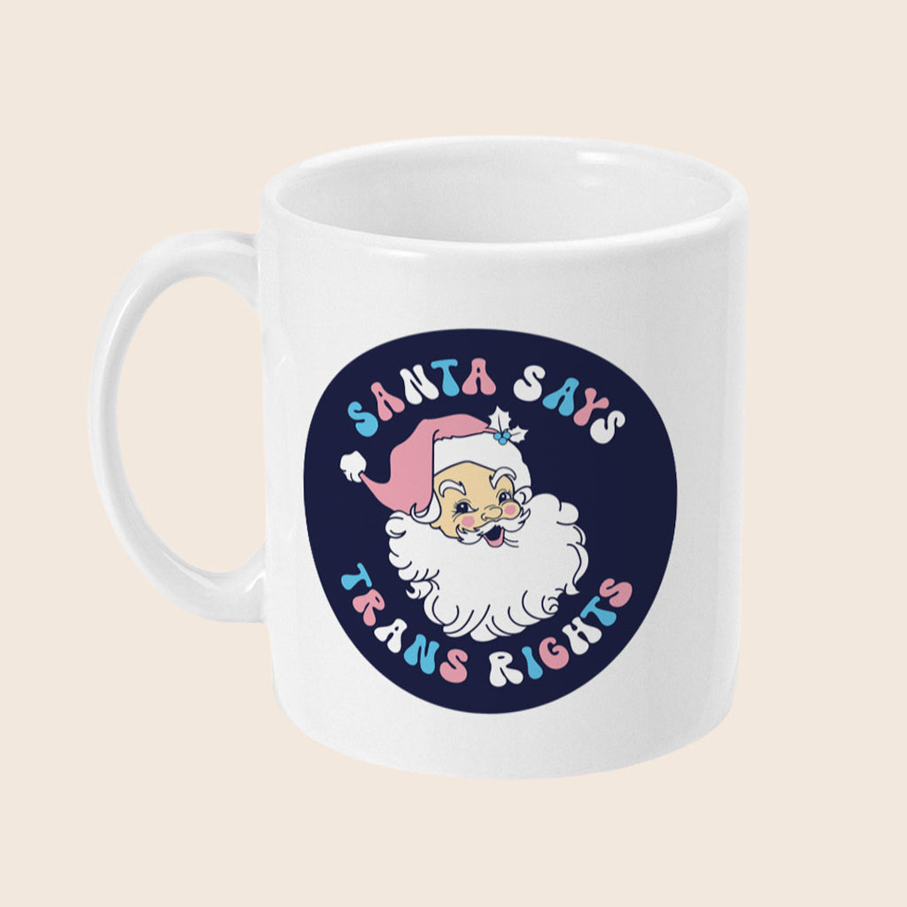White ceramic mug with a dark navy circle featuring a vintage-style Santa illustration wearing a pink hat. Surrounding Santa is pastel bubble text that reads: “Santa Says Trans Rights.”