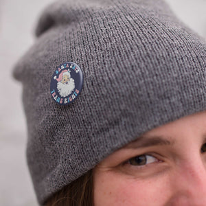 Close up of a woman wearing a grey knitted hat with a 38mm 'Santa Says Trans Rights' badge pinned to it.