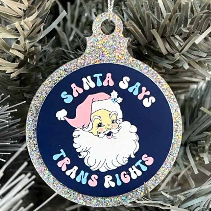 Close up of a white Christmas tree displaying a silver glitter bauble featuring the text Santa Says Trans Rights and a illustration of Santa Claus