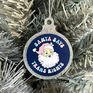 Close up of a white Christmas tree displaying a silver glitter bauble featuring the text Santa Says Trans Rights and a illustration of Santa Claus