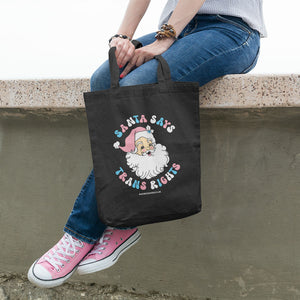 A Young Woman Sitting on a Wall holding a black tote bag with the slogan Santa Says Trans Rights with an image of Santa Claus. The text is in the colours of the transgender flag; pink, blue, and white.