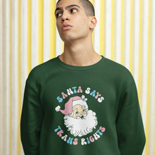 Load image into Gallery viewer, Young man wearing a bottle green sweatshirt with a Santa Says Trans Rights motif in the colours of the Transgender flag