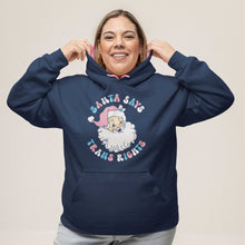 Load image into Gallery viewer, A smiling woman with shoulder length hair and hooped earrings wearing an Oxford Blue hooded sweatshirt with contrasting pink inner hood and drawcords on a white background. The hoodie features the design Santa Says Trans Rights and an image of Santa Claus in the colours of the transgender flag.