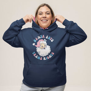 A smiling woman with shoulder length hair and hooped earrings wearing an Oxford Blue hooded sweatshirt with contrasting pink inner hood and drawcords on a white background. The hoodie features the design Santa Says Trans Rights and an image of Santa Claus in the colours of the transgender flag.