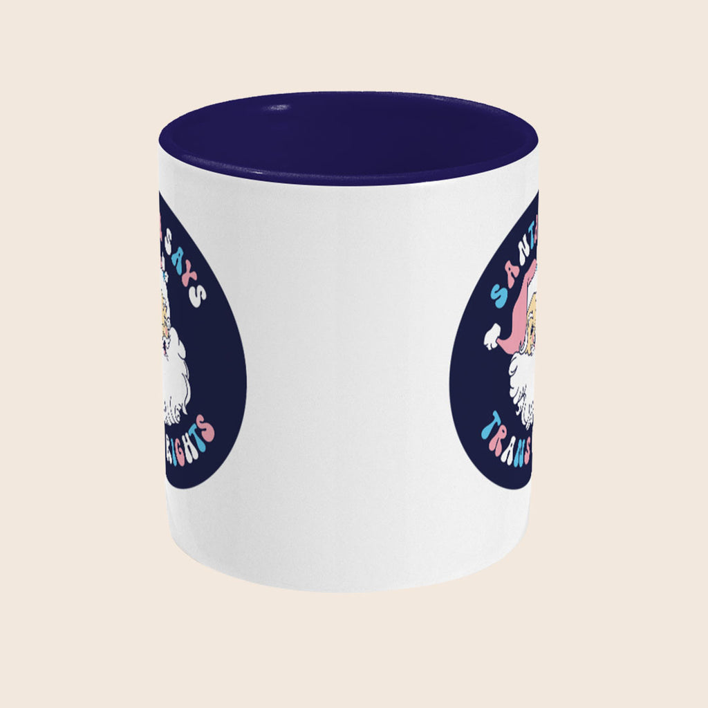 A white ceramic mug with a dark navy interior. The design features a vintage-style Santa Claus illustration wearing a pink hat, with the phrase “Santa Says Trans Rights” in pastel blue and pink letters surrounding the image.