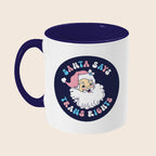 A white ceramic mug with a navy blue handle and interior. The design features a retro-style Santa Claus illustration and the phrase “Santa Says Trans Rights” in pastel tones, with the text curving around the image.