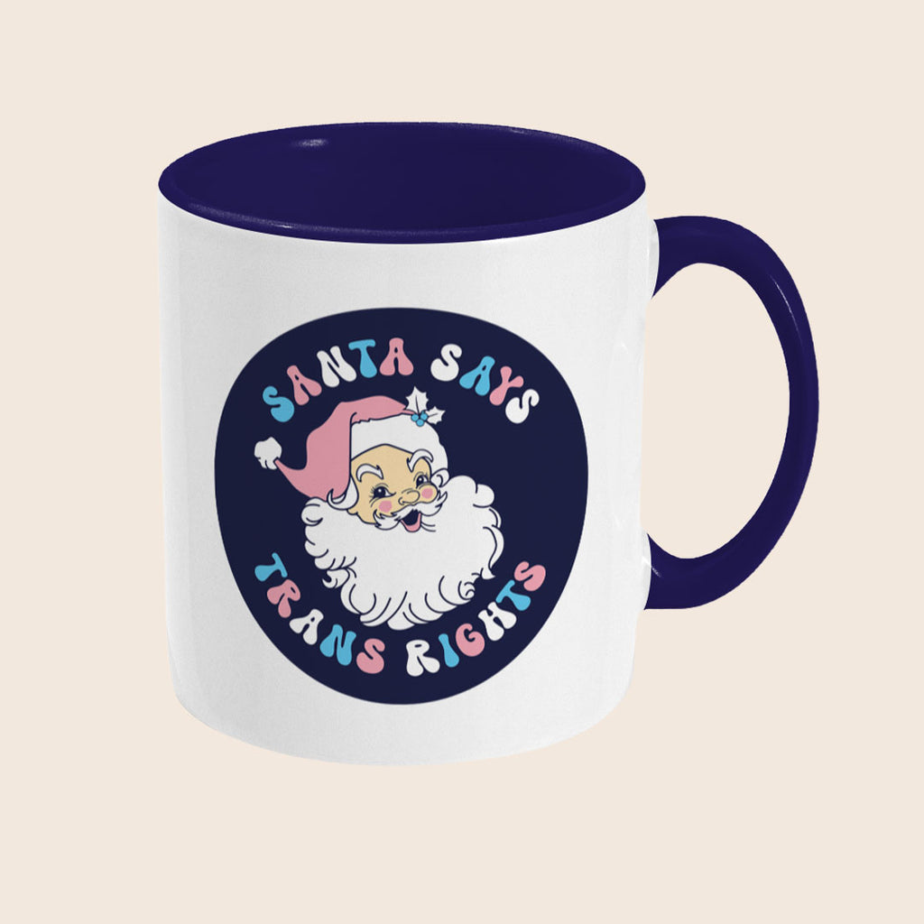 A white ceramic mug with a navy handle and interior. The front features a cheerful illustration of Santa Claus with a pink hat, surrounded by pastel lettering that reads “Santa Says Trans Rights.”