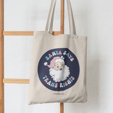 Load image into Gallery viewer, Natural cotton tote bag hanging from a wooden stair featuring the slogan Santa Says Trans Rights with an image of Santa Claus on a navy blue circle. The text is in the colours of the transgender flag; pink, blue, and white.
