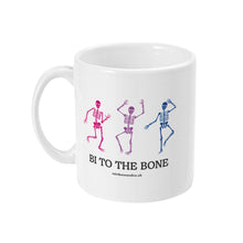 Load image into Gallery viewer, A white ceramic coffee mug with the handle on the left. The mug shows three dancing skeletons in the colours of the bisexual flag and the slogan &#39;Bi To The Bone&#39;