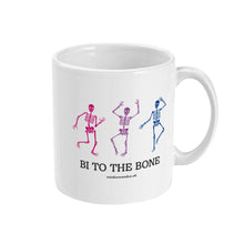Load image into Gallery viewer, A white ceramic coffee mug with the handle on the right. The mug shows three dancing skeletons in the colours of the bisexual flag and the slogan &#39;Bi To The Bone&#39;