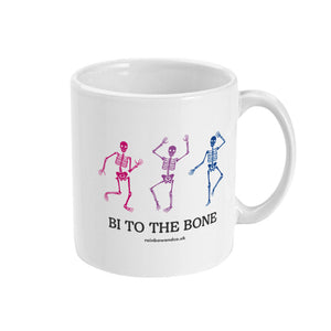 A white ceramic coffee mug with the handle on the right. The mug shows three dancing skeletons in the colours of the bisexual flag and the slogan 'Bi To The Bone'