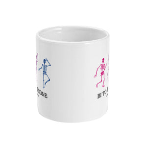 A white ceramic coffee mug with handle facing away from view. The mug shows three dancing skeletons in the colours of the bisexual flag and the slogan 'Bi To The Bone'