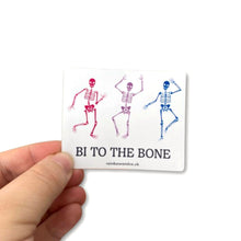 Load image into Gallery viewer, Gloss sticker being held between a thumb and forefinger. The sticker features three dancing skeletons in the colours of the bisexual flag and the slogan &#39;Bi To The Bone&#39;