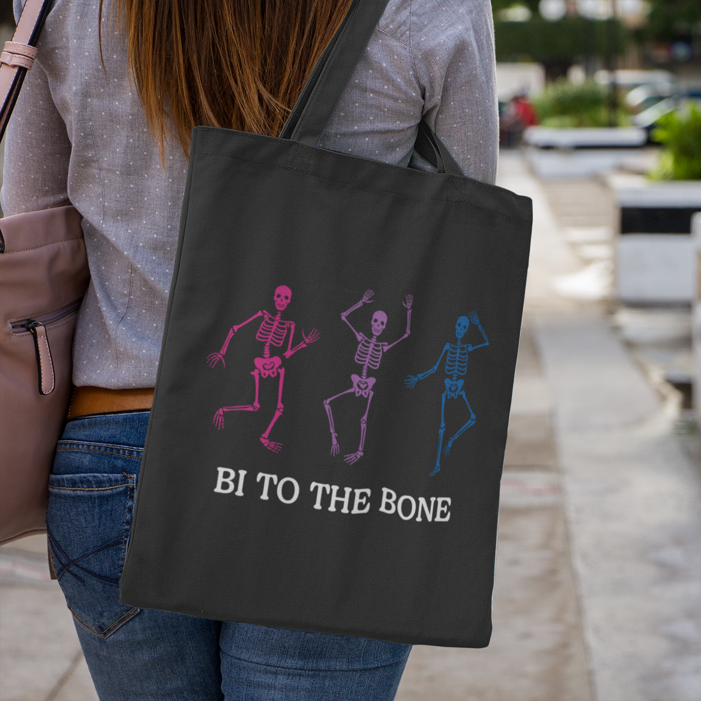 A black canvas tote bag worn over the shoulder of a woman in jeans and a spotted shirt. The bag features three dancing skeletons in pink, purple, and blue with the phrase “Bi To The Bone” in bold white text below.