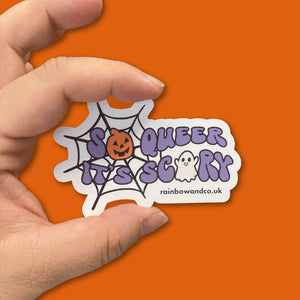 Glossy sticker featuring the phrase 'So Queer It's Scary' being held between a persons thumb and forefinger against an orange background.