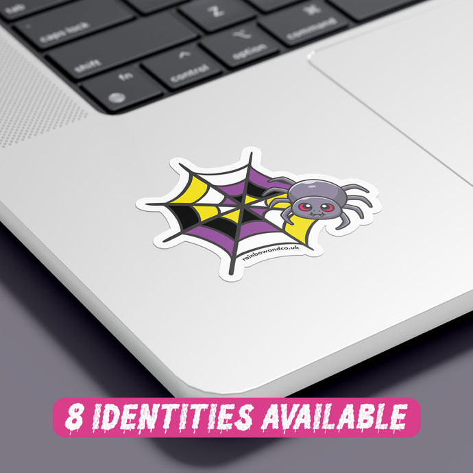 A colourful sticker featuring a cartoon spider with red eyes, resting on a spider web with a pattern that combines black, yellow, white, and purple colours, representing the non binary flag. The sticker is placed on the corner of a silver laptop, showcasing its vibrant design against the sleek surface. At the bottom, there is text in a playful pink font that reads 