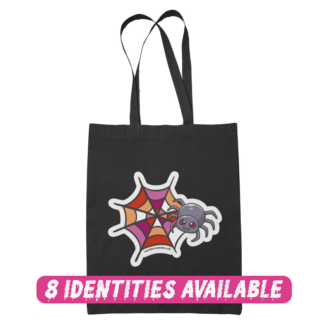 A black tote bag featuring a cute cartoon spider in front of a spider web coloured in the lesbian pride flag colours: pink, white, and orange. The text "8 IDENTITIES AVAILABLE" is written below in bold pink lettering.