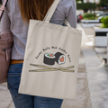 A natural canvas tote bag worn over the shoulder of a woman walking through a paved urban area. The design shows two sushi rolls and crossed chopsticks, with the phrase “Sushi Rolls Not Gender Roles” curved above.