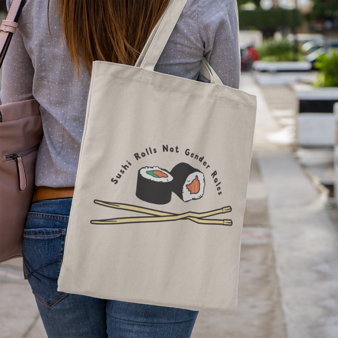 A natural canvas tote bag worn over the shoulder of a woman walking through a paved urban area. The design shows two sushi rolls and crossed chopsticks, with the phrase “Sushi Rolls Not Gender Roles” curved above.