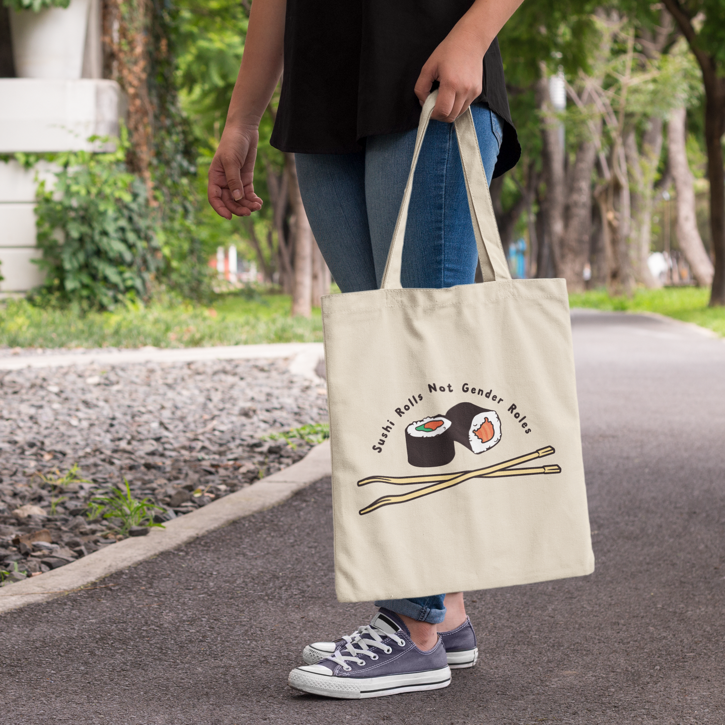 A natural canvas tote bag carried by someone walking along a tree-lined path. The bag features a playful sushi design with chopsticks and the slogan “Sushi Rolls Not Gender Roles” above.