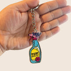 A close-up of a hand holding the TERF Repellent keyring. The design features a colourful spray bottle with "TERF Repellent – 100% Effective" written on the front. The Rainbow & Co charm is visible near the keyring loop.