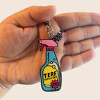 A hand holding the full TERF Repellent keyring, showing the entire silver chain, pink spray top, and attached Rainbow & Co charm. The background is a soft cream colour, contrasting with the bright design of the keyring.