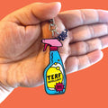 A hand holds a vibrant acrylic keyring shaped like a spray bottle against a bold orange background. The bottle reads “TERF REPELLENT” in black text over a yellow liquid design, with a pink burst label that says “100% EFFECTIVE.” The spray top is pink with a black outline, and a small clear charm displaying the Rainbow & Co logo hangs from the keyring chain.