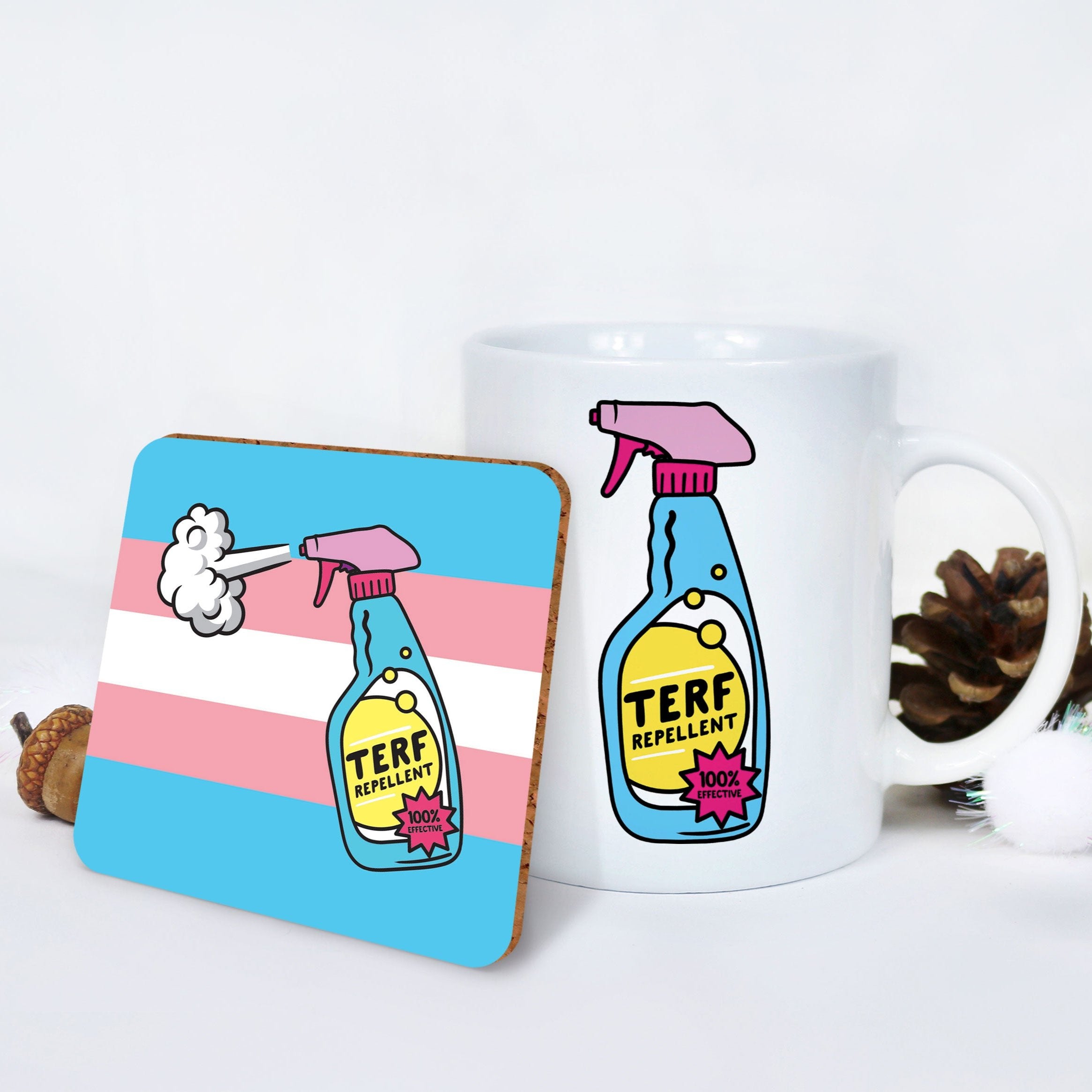 A cosy holiday setting with a white mug displaying the "TERF Repellent" spray bottle design, paired with a matching coaster over the transgender pride flag. Surrounded by pinecones and fluffy decorations, it's perfect for gifting.
