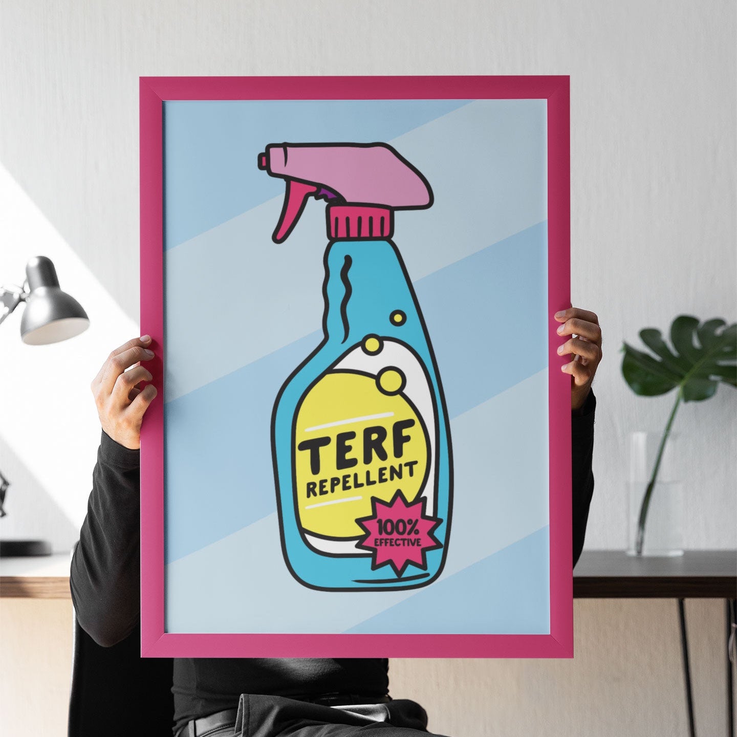 A person seated on a black chair holds up a large framed print in front of their face. The print features a bold, cartoon-style spray bottle labelled "TERF REPELLENT" with the phrase “100% EFFECTIVE” in a pink starburst. The bottle is bright blue with yellow liquid and a pink nozzle, set against a soft blue striped background. The frame is a vibrant pink, and the setting appears to be a modern home or office with a plant and lamp in the background.