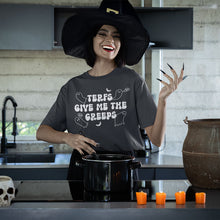 Load image into Gallery viewer, A woman wearing a dark grey &quot;TERFs Give Me The Creeps&quot; T-shirt stands in a kitchen dressed in a witch&#39;s hat, with long black nails and bright red lipstick. She is smiling while stirring a pot on the stove, surrounded by Halloween decorations like a skull, an old cauldron, and orange candles, adding a spooky Halloween vibe to the scene.