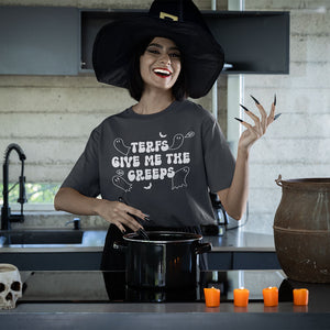 A woman wearing a dark grey "TERFs Give Me The Creeps" T-shirt stands in a kitchen dressed in a witch's hat, with long black nails and bright red lipstick. She is smiling while stirring a pot on the stove, surrounded by Halloween decorations like a skull, an old cauldron, and orange candles, adding a spooky Halloween vibe to the scene.