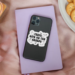 A white sticker with the text "TERFs Give Me The Creeps" in black, surrounded by small ghost and bat illustrations, is applied to the back of a black smartphone case. The phone rests on a light purple notebook placed on a soft pink fabric surface next to a lit candle and a plate of crackers.