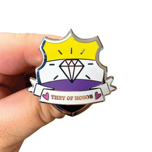 Load image into Gallery viewer, White hand holding up an enamel pin in the shape of a shield with a banner across it&#39;s front. Banner reads They of Honor. The background of the pin is the non binary pride flag and there is a jewel icon in the centre. The jewel and banner are filled with white glitter.