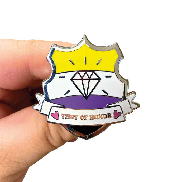 White hand holding up an enamel pin in the shape of a shield with a banner across it's front. Banner reads They of Honor. The background of the pin is the non binary pride flag and there is a jewel icon in the centre. The jewel and banner are filled with white glitter.