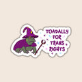 A die-cut sticker featuring a cartoon green toad wearing a purple witch hat and cape, holding a wand, next to the text 'TOADALLY FOR TRANS RIGHTS' in purple with small star accents, shown on a light beige background.