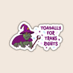 A die-cut sticker featuring a cartoon green toad wearing a purple witch hat and cape, holding a wand, next to the text 'TOADALLY FOR TRANS RIGHTS' in purple with small star accents, shown on a light beige background.