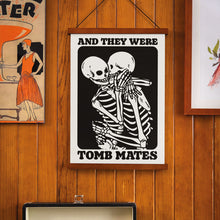 Load image into Gallery viewer, A framed poster titled &quot;And They Were Tomb Mates&quot; featuring two skeletons in an embrace. The print is displayed on a wooden wall, with other vintage-style artworks, including a woman in an orange dress next to a drink, providing a retro vibe. The poster hangs from a wooden frame with twine, complementing the rustic decor.