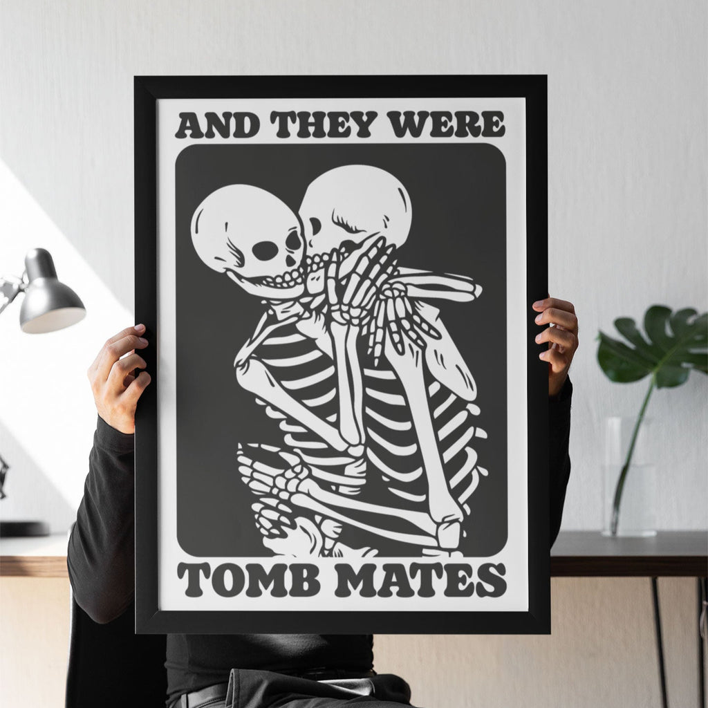A person sits holding a black-and-white framed print featuring two skeletons kissing with the phrase "And They Were Tomb Mates," styled in a gothic, vintage-inspired design.