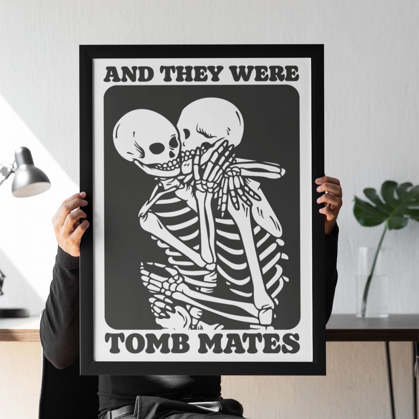 A person sits holding a black-and-white framed print featuring two skeletons kissing with the phrase "And They Were Tomb Mates," styled in a gothic, vintage-inspired design.