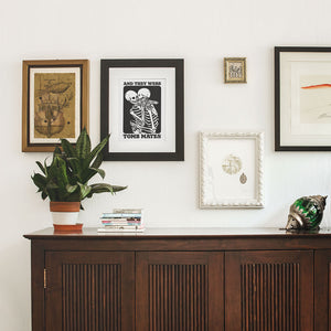 A framed print titled "And They Were Tomb Mates" featuring two skeletons in an embrace, hung on a white wall among other artwork. The skeletons are depicted with one leaning on the other's shoulder. The print is in black and white with bold text, surrounded by additional framed artworks, a green potted plant on a wooden cabinet, and a small stack of books.