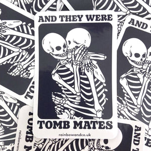 A pile of glossy stickers with one sticker in the foreground. The stickers show two skeletons embracing and the text 'And They Were Tomb Mates'
