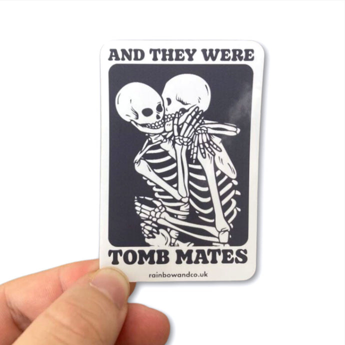 Gloss sticker being held between the thumb and forefinger. The sticker shows two skeletons embracing and the text 'And They Were Tomb Mates'