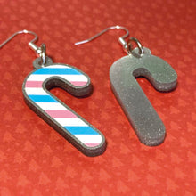 Load image into Gallery viewer, Silver glitter candy cane dangle earrings lay on a red christmas tree paper background. The stripes on the candy cane are those of the Transgender flag; blue, pink, and white. One of the earrings is turned over to show the reverse, which is plain silver glitter.