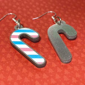 Silver glitter candy cane dangle earrings lay on a red christmas tree paper background. The stripes on the candy cane are those of the Transgender flag; blue, pink, and white. One of the earrings is turned over to show the reverse, which is plain silver glitter.