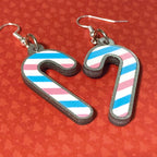 Silver glitter candy cane dangle earrings lay on a red christmas tree paper background. The stripes on the candy cane are those of the Transgender flag; blue, pink, white. One of the earrings is turned over to show the reverse, which is plain silver glitter.