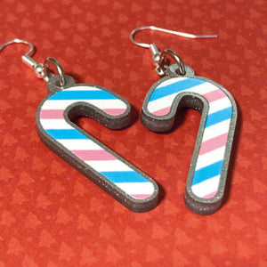 Silver glitter candy cane dangle earrings lay on a red christmas tree paper background. The stripes on the candy cane are those of the Transgender flag; blue, pink, white. One of the earrings is turned over to show the reverse, which is plain silver glitter.