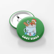Load image into Gallery viewer, Close up of a green 38mm badge featuring retro text reading &#39;cosy vibes&#39;. The image shows a mug of hot chocolate with marshmallows and a gingerbread man wearing a Santa hat. A candy cane in the mug and lights around the mug are the colours of the transgender flag.