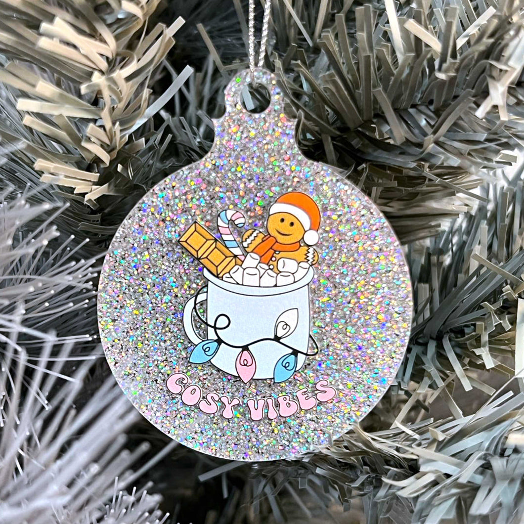 Close up of a white Christmas tree displaying a silver glitter bauble featuring retro text reading 'cosy vibes'. The image shows a mug of hot chocolate with marshmallows and a gingerbread man wearing a Santa hat. A candy cane in the mug and lights around the mug are the colours of the transgender pride flag.